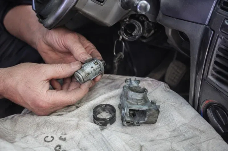 Hands holding a removed ignition cylinder beside a car steering column, with the housing and small parts on a cloth. — Ignition Lock Repair, King